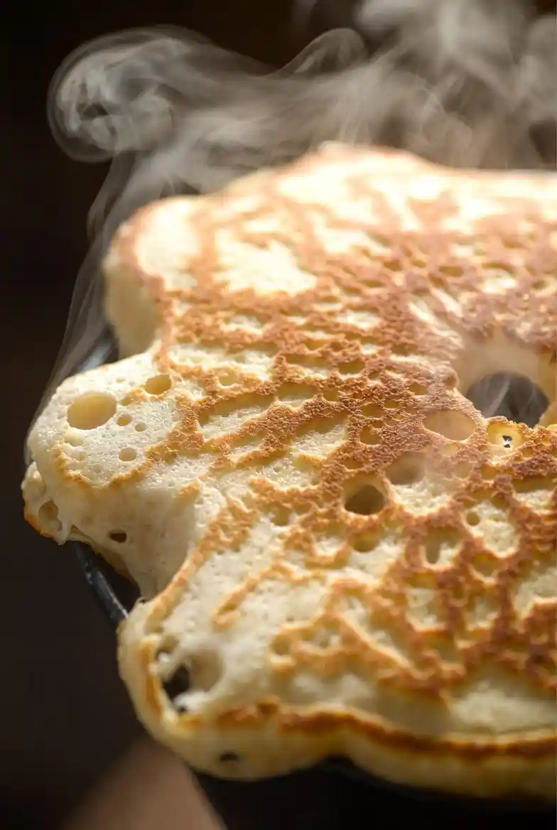 Extreme close-up of a fluffy pancake surface showing soft, airy texture with tiny bubbles and slight steam rising, highlighting a warm golden, freshly cooked appearance.
