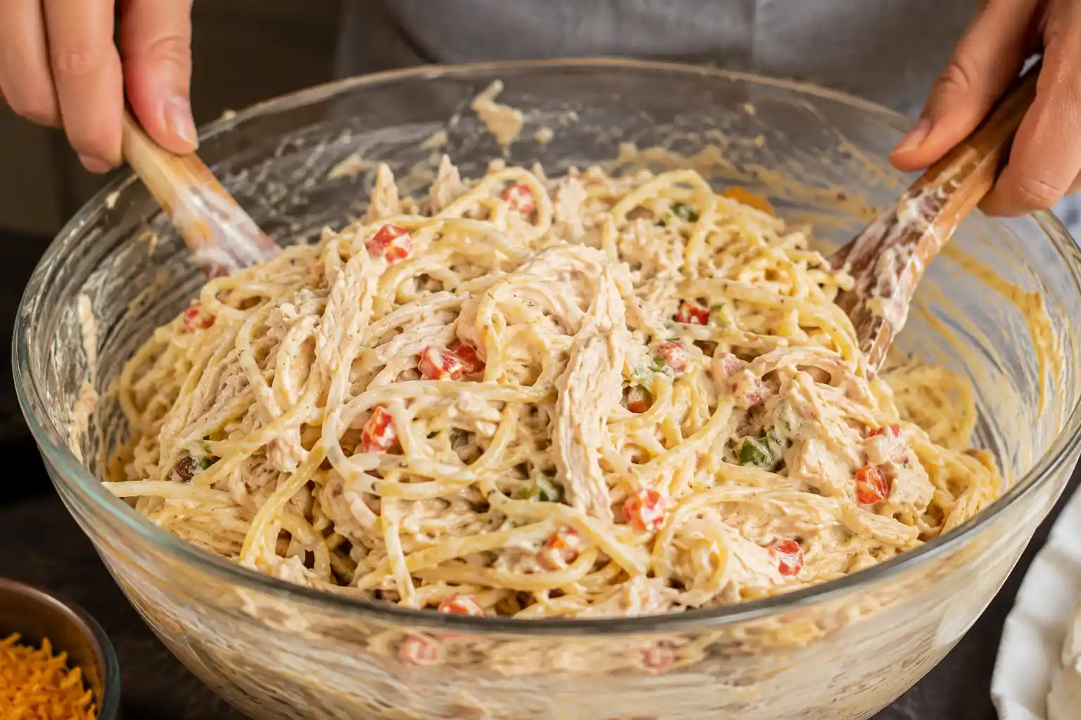Hands mixing spaghetti, shredded chicken, and creamy sauce in a large bowl before baking.