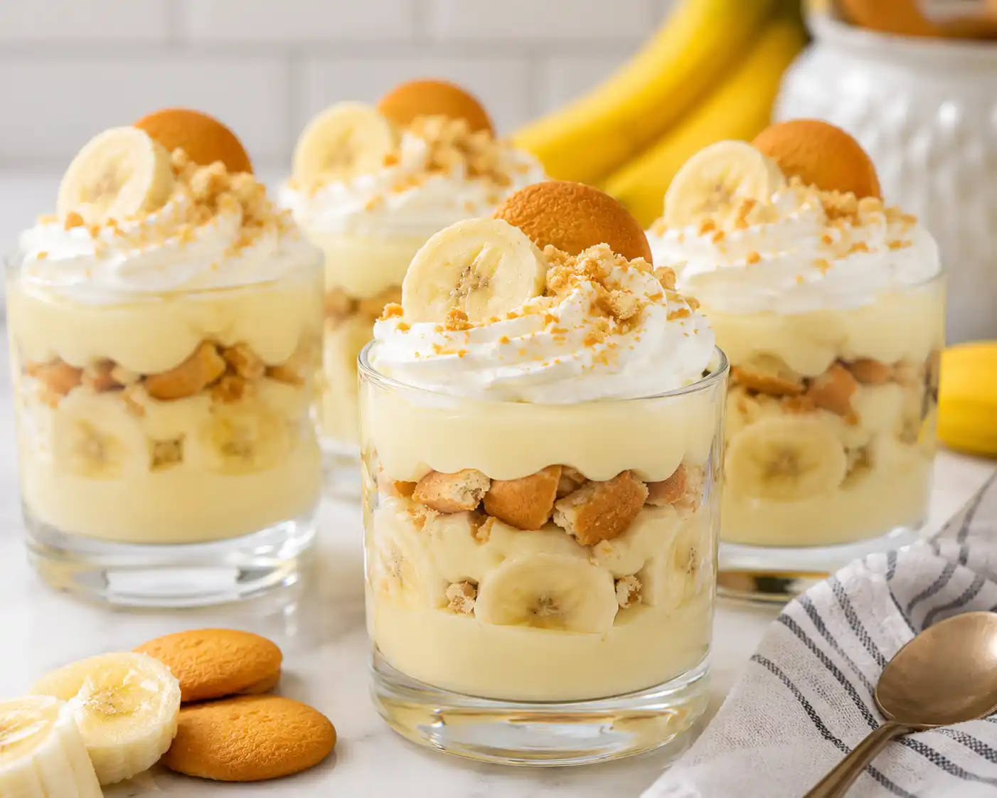 Individual glass cups filled with no bake banana pudding topped with whipped cream banana slices and crushed wafers in a clean kitchen setting